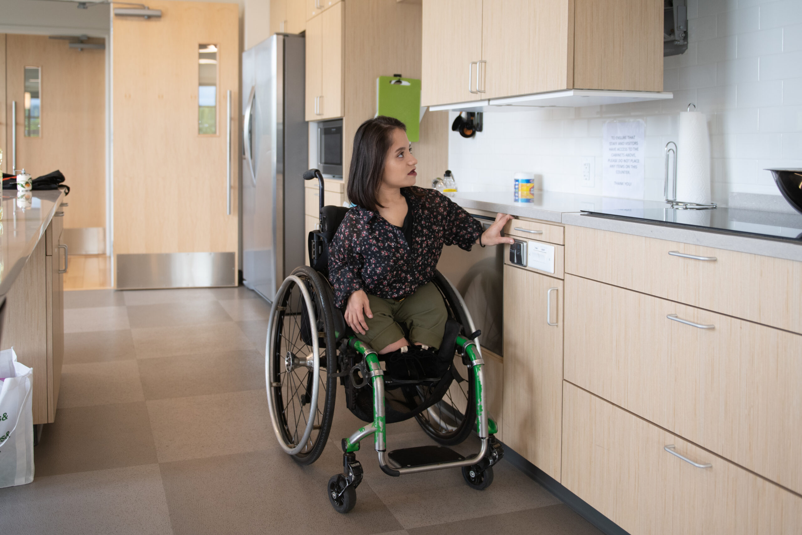 dah-kitchen01 A South Asian person sits in her wheelchair and presses a button to lower the height-adjustable shelves in an accessible kitchen.