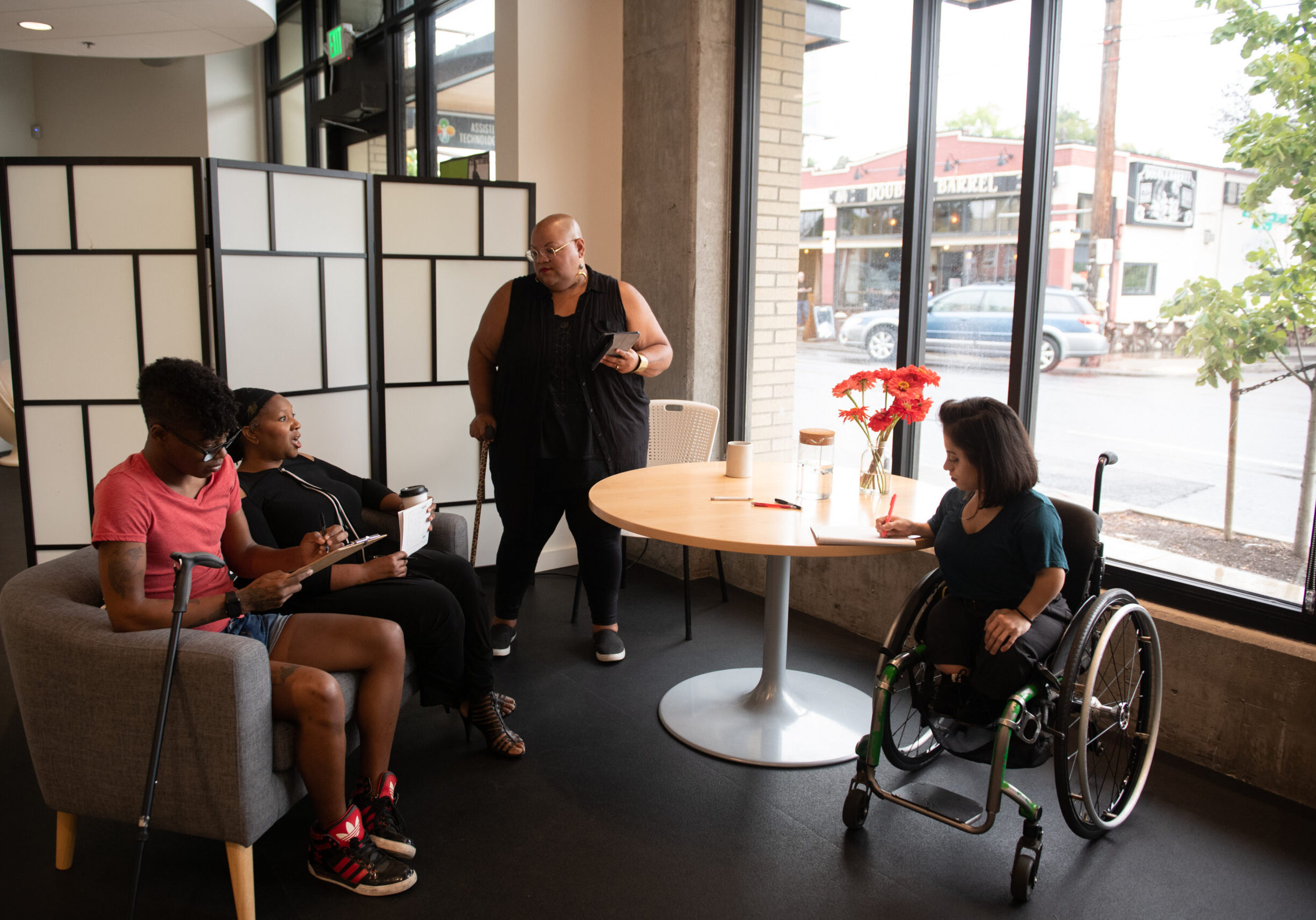 dah-meeting04 Four disabled people of color face each other in a open circle during a meeting. Two Black people sit on a couch with a cane leaning off the side while a Black non-binary person stands with a tablet and cane. A South Asian person in a wheelchair takes notes.